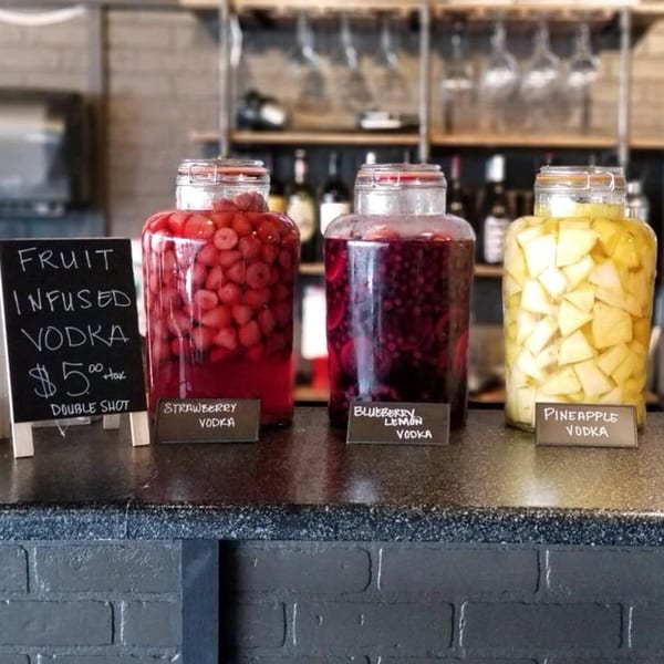 Three large jars of fruit-infused vodka (strawberry, blueberry lemon, pineapple) on a bar counter.